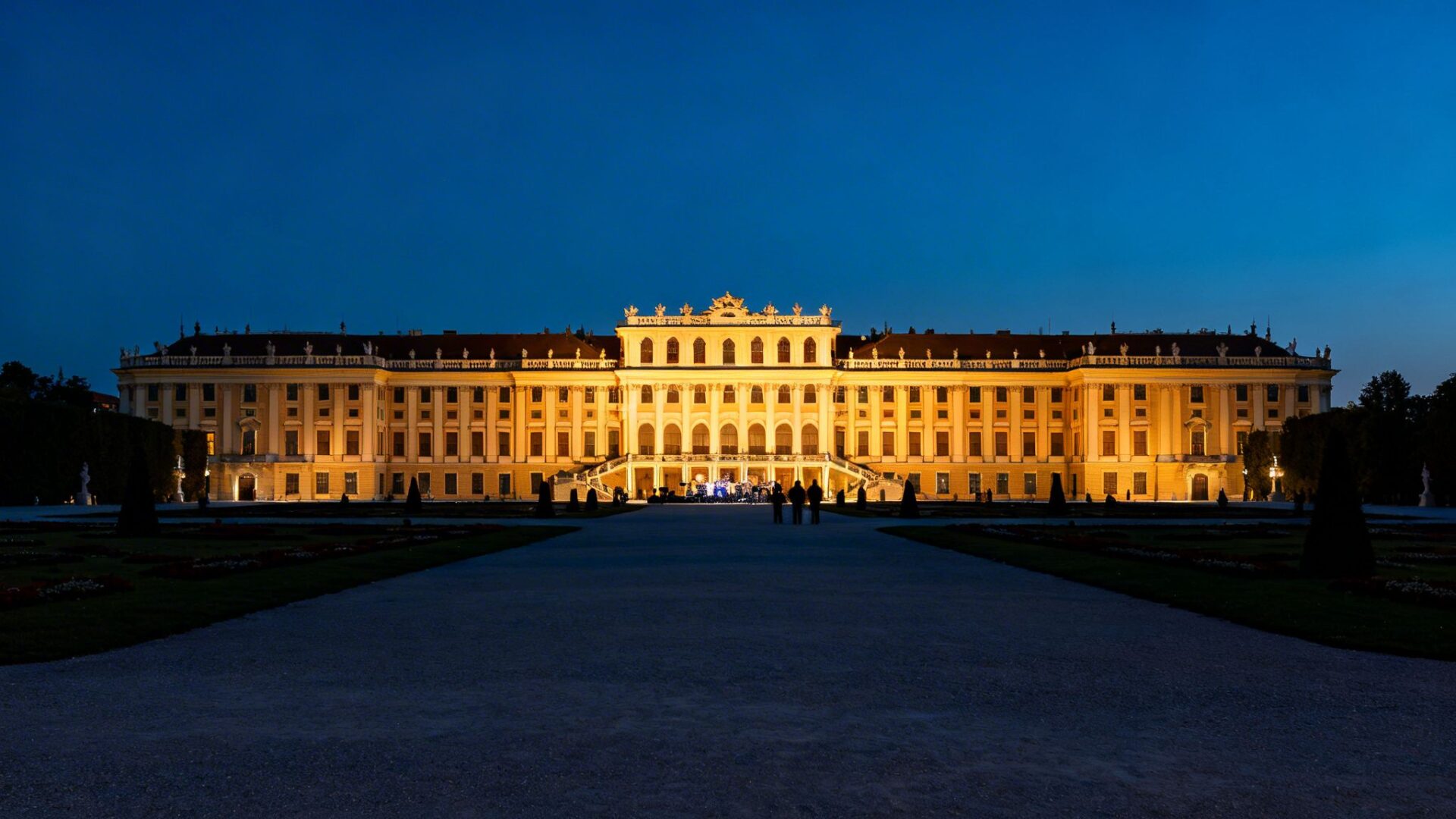 The grand Schönbrunn Palace illuminated at night, with people gathered in the garden for a concert.